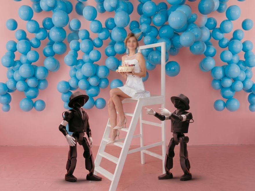 Deux robots coiffés de chapeaux qui posent à côté d'une femme tenant un gâteau de fête, devant des ballons bleus
